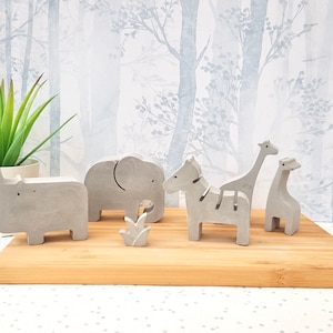 May include: A set of five grey concrete animal figurines, including a rhino, elephant, zebra, and two giraffes, displayed on a wooden surface. The figurines are stylised and have a minimalist design.
