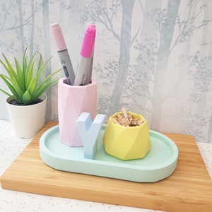 K&ouml;nnte beinhalten: Ein geometrischer Schreibtisch-Organizer in Pastellrosa, Blau und Gelb mit einem kleinen Tablett, einem Stifthalter und einem kleinen Beh&auml;lter f&uuml;r B&uuml;roklammern. Der Organizer steht auf einer Holzoberfl&auml;che.
