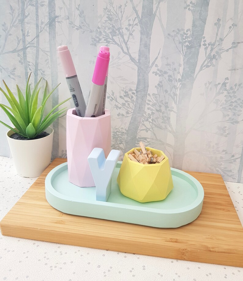 K&ouml;nnte beinhalten: Ein geometrischer Schreibtisch-Organizer in Pastellrosa, Blau und Gelb mit einem kleinen Tablett, einem Stifthalter und einem kleinen Beh&auml;lter f&uuml;r B&uuml;roklammern. Der Organizer steht auf einer Holzoberfl&auml;che.