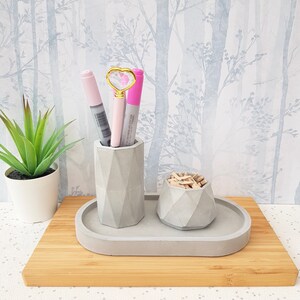 May include: A grey concrete desk organiser with two compartments. The larger compartment holds pens and the smaller compartment holds paper clips. The organiser is sitting on a wooden surface.