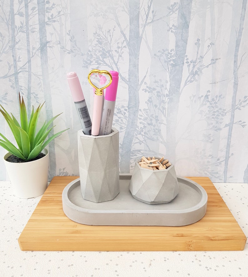 May include: A grey concrete desk organiser with two compartments. The larger compartment holds pens and the smaller compartment holds paper clips. The organiser is sitting on a wooden surface.