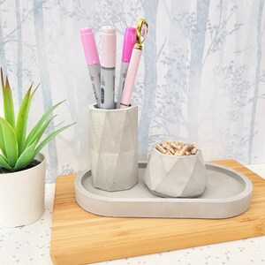 May include: A grey concrete desk organiser with three compartments. The organiser has a geometric design and is sitting on a wooden surface. The compartments hold pens, pencils, and paper clips.