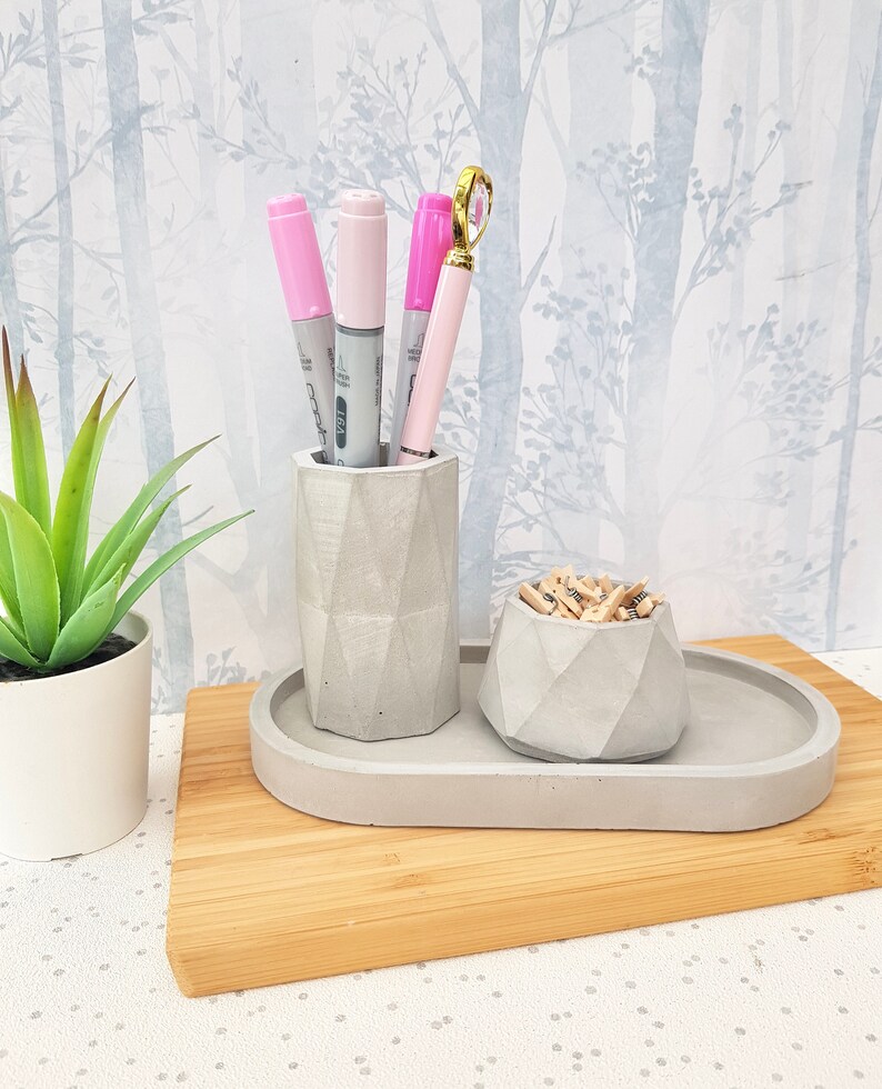 May include: A grey concrete desk organiser with three compartments. The organiser has a geometric design and is sitting on a wooden surface. The compartments hold pens, pencils, and paper clips.