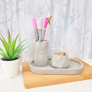 May include: A grey concrete desk organiser with three compartments. The organiser has a small bowl for paper clips and a tall cylinder for pens and pencils. The organiser is sitting on a wooden surface.