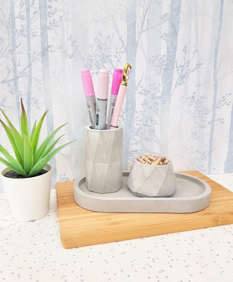 May include: A grey concrete desk organiser with three compartments. The organiser has a small bowl for paper clips and a tall cylinder for pens and pencils. The organiser is sitting on a wooden surface.