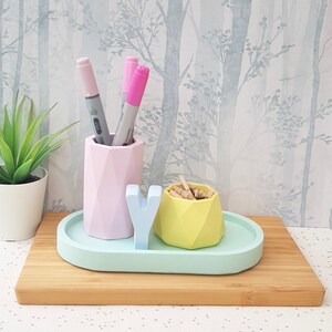 K&ouml;nnte beinhalten: Ein pastellblauer Schreibtisch-Organizer mit drei F&auml;chern. Die F&auml;cher sind aus geometrischen Formen gefertigt und halten rosa und graue Marker sowie Holzklammern. Der Organizer steht auf einer hellbraunen Holzoberfl&auml;che.