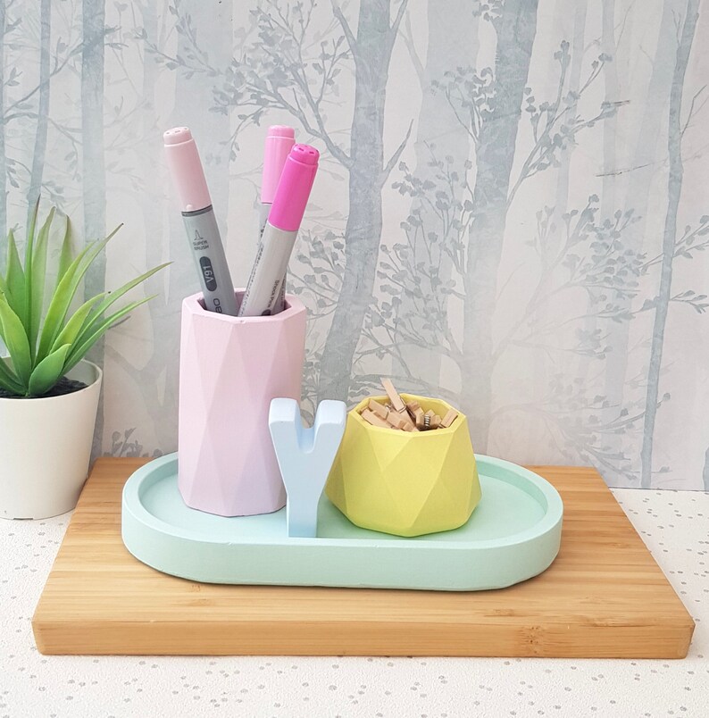 K&ouml;nnte beinhalten: Ein pastellblauer Schreibtisch-Organizer mit drei F&auml;chern. Die F&auml;cher sind aus geometrischen Formen gefertigt und halten rosa und graue Marker sowie Holzklammern. Der Organizer steht auf einer hellbraunen Holzoberfl&auml;che.