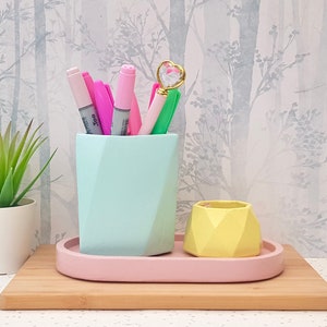 May include: A light blue geometric ceramic pencil holder with pink and green markers and a pink heart-shaped wand inside. The holder is sitting on a pink tray on a wooden surface.