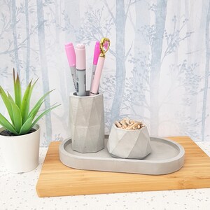 May include: A grey concrete desk organiser with a pen holder and a small bowl for paper clips. The organiser is on a wooden surface.