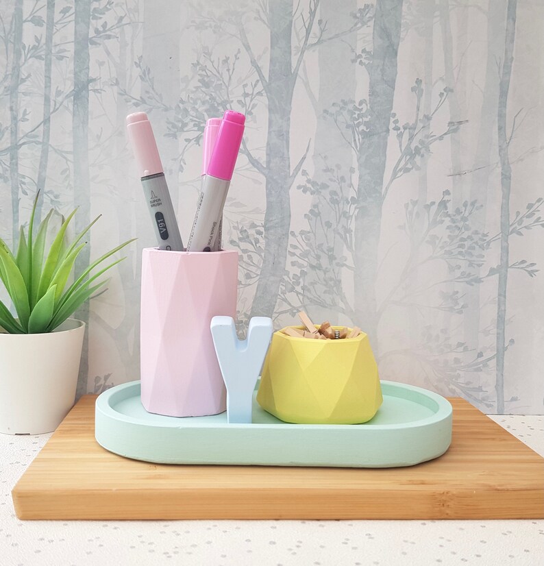 K&ouml;nnte beinhalten: Ein pastellgr&uuml;nes ovales Tablett mit einem geometrischen Keramikhalter in Rosa und Gelb f&uuml;r Stifte und Bleistifte, einem geometrischen Keramikhalter in Gelb f&uuml;r B&uuml;roklammern und einem gro&szlig;en wei&szlig;en Buchstaben "Y" auf dem Tablett.