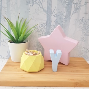May include: A yellow geometric container with wooden clothes pegs, a light blue wooden letter Y, and a pink star-shaped ornament on a wooden surface.