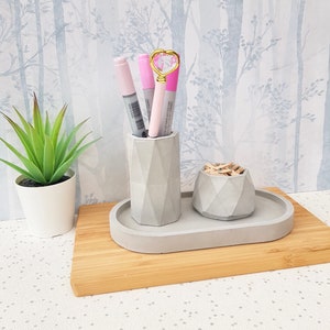 May include: A grey concrete desk organiser with three compartments. The organiser has a small round compartment for paper clips, a larger round compartment for pens and pencils, and a rectangular tray for other items. The organiser is sitting on a wooden surface.