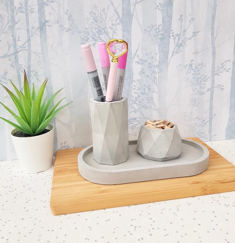May include: A grey concrete desk organiser with three compartments. The organiser has a small round compartment for paper clips, a larger round compartment for pens and pencils, and a rectangular tray for other items. The organiser is sitting on a wooden surface.