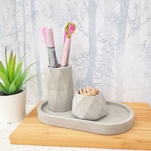 May include: A grey concrete desk organiser with three compartments. The organiser has a rectangular tray, a tall cylindrical container, and a smaller round container. The tray is empty, the cylinder holds three pink pens, and the round container holds wooden push pins.
