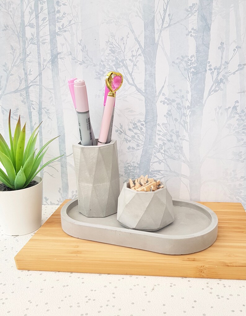 May include: A grey concrete desk organiser with three compartments. The organiser has a rectangular tray, a tall cylindrical container, and a smaller round container. The tray is empty, the cylinder holds three pink pens, and the round container holds wooden push pins.