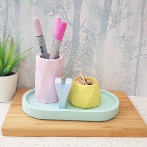 K&ouml;nnte beinhalten: Ein pastellblaues Keramiktablett mit einem rosa geometrischen Stifthalter, einem gelben geometrischen Beh&auml;lter und einem wei&szlig;en Buchstaben 'Y'. Das Tablett befindet sich auf einer Holzoberfl&auml;che.