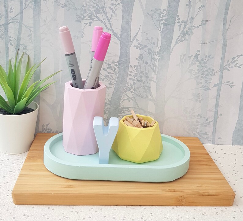 K&ouml;nnte beinhalten: Ein pastellblaues Keramiktablett mit einem rosa geometrischen Stifthalter, einem gelben geometrischen Beh&auml;lter und einem wei&szlig;en Buchstaben 'Y'. Das Tablett befindet sich auf einer Holzoberfl&auml;che.