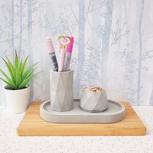 May include: A grey concrete desk organiser with three compartments. The organiser has a geometric design and is sitting on a wooden surface. There are three pens and a heart-shaped paperclip in the organiser.