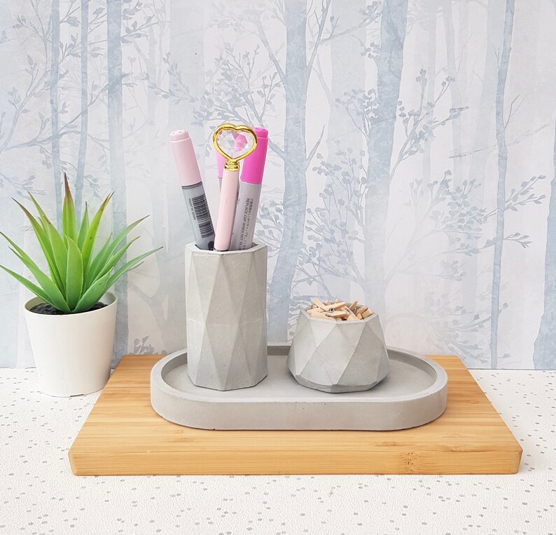 May include: A grey concrete desk organiser with three compartments. The organiser has a geometric design and is sitting on a wooden surface. There are three pens and a heart-shaped paperclip in the organiser.