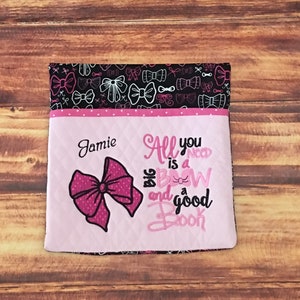 May include: Pink and black book bag with a pink bow and the text "All you need is a big bow and a good book" embroidered on the front. The bag is personalized with the name "Jamie".