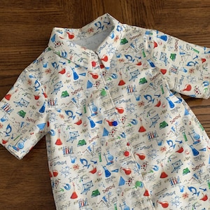 May include: A short-sleeved button-down shirt with a white background and a colorful science-themed pattern. The pattern includes beakers, molecules, and other scientific symbols in red, blue, and green. The shirt has a classic collar.