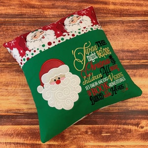 K&ouml;nnte beinhalten: Ein weihnachtliches Kissen mit einem gr&uuml;nen Mittelteil und einem roten Rand mit Weihnachtsmann-Gesichtern und Schneeflocken. Das Kissen hat ein gesticktes Weihnachtsmann-Gesicht und den Text "Twas the night before Christmas..."