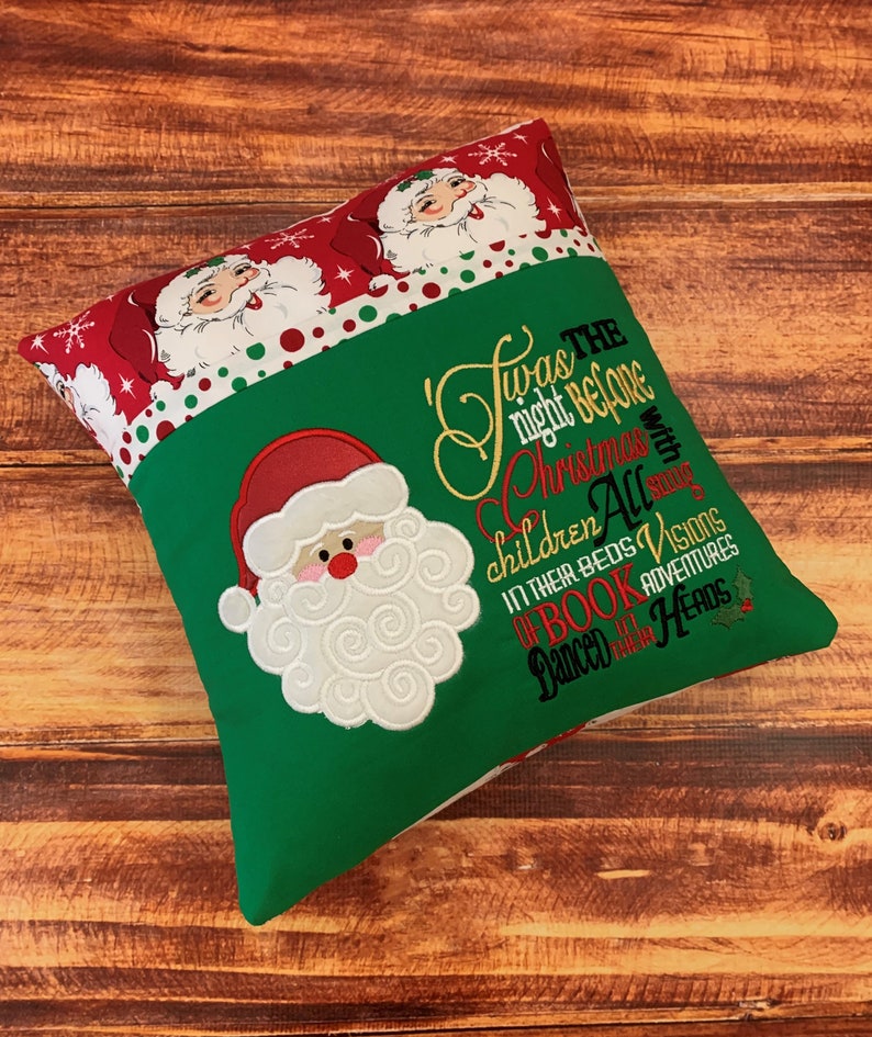 K&ouml;nnte beinhalten: Ein weihnachtliches Kissen mit einem gr&uuml;nen Mittelteil und einem roten Rand mit Weihnachtsmann-Gesichtern und Schneeflocken. Das Kissen hat ein gesticktes Weihnachtsmann-Gesicht und den Text "Twas the night before Christmas..."