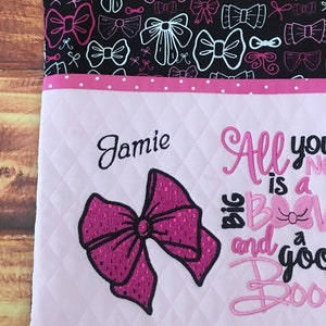 May include: A pink and white quilted fabric bag with a pink bow and the text "All you need is a big bow and a good boo boo". The bag has a black fabric lining with a white bow pattern. The name "Jamie" is embroidered on the front of the bag.
