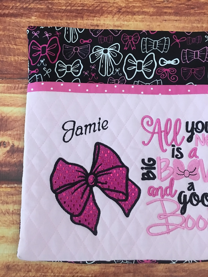 May include: A pink and white quilted fabric bag with a pink bow and the text "All you need is a big bow and a good boo boo". The bag has a black fabric lining with a white bow pattern. The name "Jamie" is embroidered on the front of the bag.