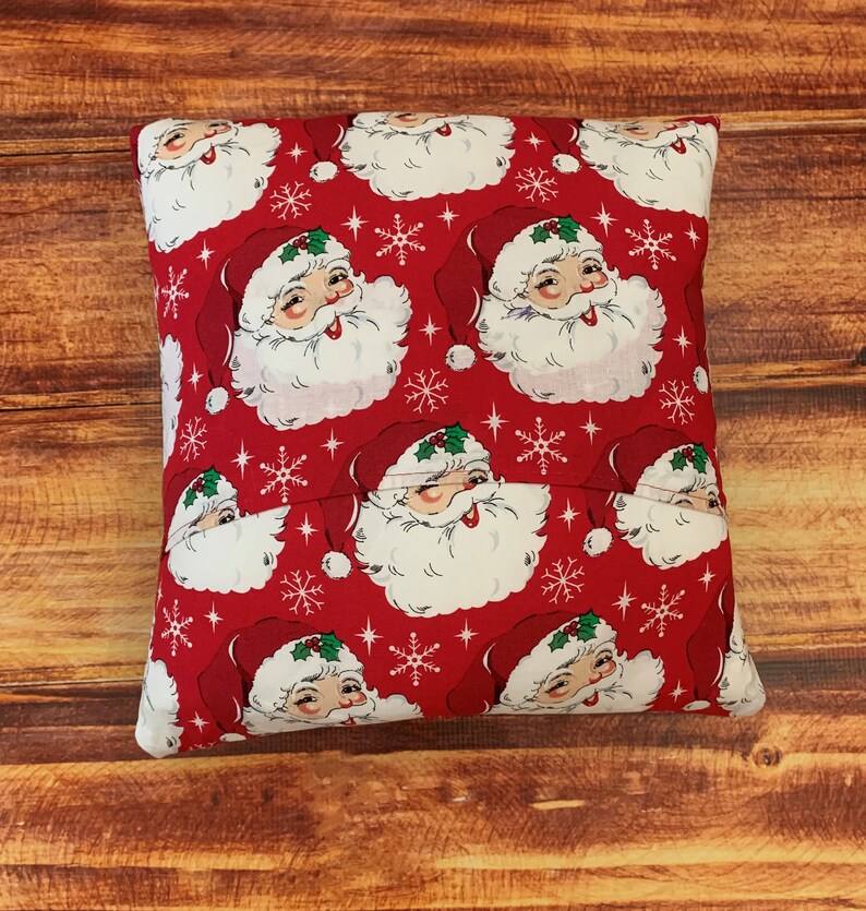 K&ouml;nnte beinhalten: Ein quadratisches, rotes Weihnachtskissen mit einem sich wiederholenden Muster von Weihnachtsmann-Gesichtern mit wei&szlig;en B&auml;rten und roten H&uuml;ten. Wei&szlig;e Schneeflocken und Sterne sind &uuml;ber das Design verstreut. Das Kissen liegt auf einer Holzoberfl&auml;che.