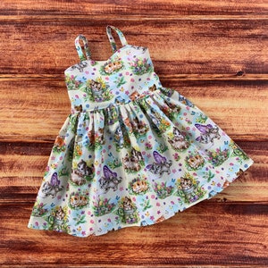 Pu&ograve; includere: Un abito senza maniche in tessuto chiaro con un motivo ripetuto di gattini, alcuni con ali di farfalla viola, immersi tra fiori colorati, fogliame verde e annaffiatoi, realizzato con sottili spalline sulle spalle.