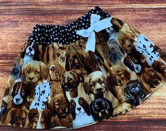Può includere: Una gonna marrone e bianca con un motivo di volti di cani. La gonna ha una cintura a pois neri e bianchi e un fiocco di raso bianco.
