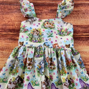 K&ouml;nnte beinhalten: Ein &auml;rmelloses Kleid mit hellcremefarbenem Hintergrund und einem wiederkehrenden Muster aus K&auml;tzchen, bunten Blumen, Schmetterlingen und Vogelh&auml;uschen. Das Oberteil hat ger&uuml;schte Tr&auml;ger und einen gerafften Rock mit weiter, plissierter Form.