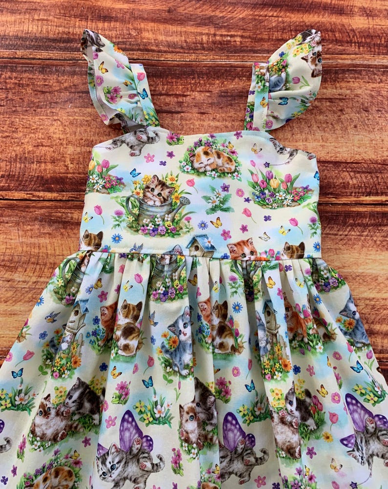 K&ouml;nnte beinhalten: Ein &auml;rmelloses Kleid mit hellcremefarbenem Hintergrund und einem wiederkehrenden Muster aus K&auml;tzchen, bunten Blumen, Schmetterlingen und Vogelh&auml;uschen. Das Oberteil hat ger&uuml;schte Tr&auml;ger und einen gerafften Rock mit weiter, plissierter Form.