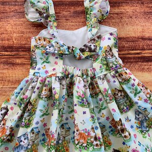 K&ouml;nnte beinhalten: Ein &auml;rmelloses Kleid aus hellem Stoff mit einem Allover-Druck von K&auml;tzchen, Vogelh&auml;uschen, bunten Blumen und Schmetterlingen. Das Kleid hat ger&uuml;schte Schultertr&auml;ger und ein Bindedetail am oberen R&uuml;cken mit einem Ausschnitt.