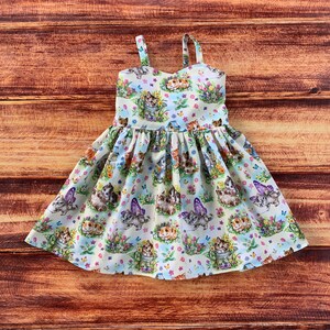 Pu&ograve; includere: Un abito senza maniche in tessuto chiaro con una stampa all-over di gattini, farfalle e fiori colorati. L'abito presenta un corpetto aderente con spalline sottili e una gonna arricciata e svasata che arriva fino alle ginocchia.