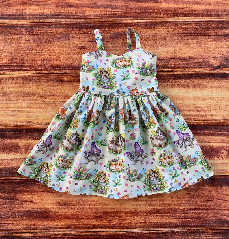 Pu&ograve; includere: Un abito senza maniche in tessuto chiaro con una stampa all-over di gattini, farfalle e fiori colorati. L'abito presenta un corpetto aderente con spalline sottili e una gonna arricciata e svasata che arriva fino alle ginocchia.