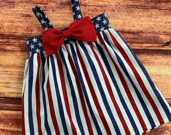 May include: A red, white, and blue striped dress with a large red bow. The dress has a sleeveless design and a gathered skirt.