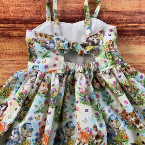 Pu&ograve; includere: Un abito senza maniche in tessuto chiaro con un motivo ripetuto di gattini, casette per uccelli, farfalle e fiori colorati. L'abito presenta spalline sottili, un dettaglio cut-out sulla schiena con un laccio e una gonna arricciata.
