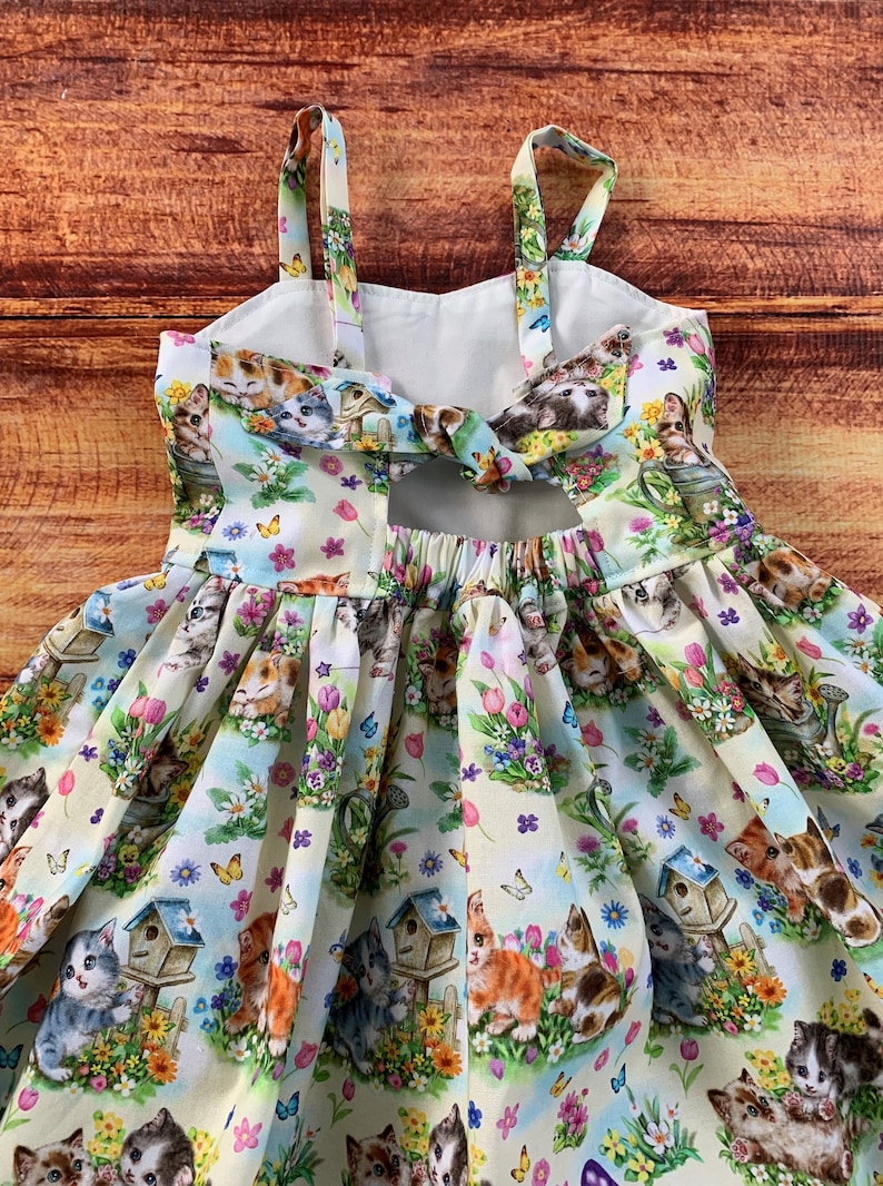 Pu&ograve; includere: Un abito senza maniche in tessuto chiaro con un motivo ripetuto di gattini, casette per uccelli, farfalle e fiori colorati. L'abito presenta spalline sottili, un dettaglio cut-out sulla schiena con un laccio e una gonna arricciata.