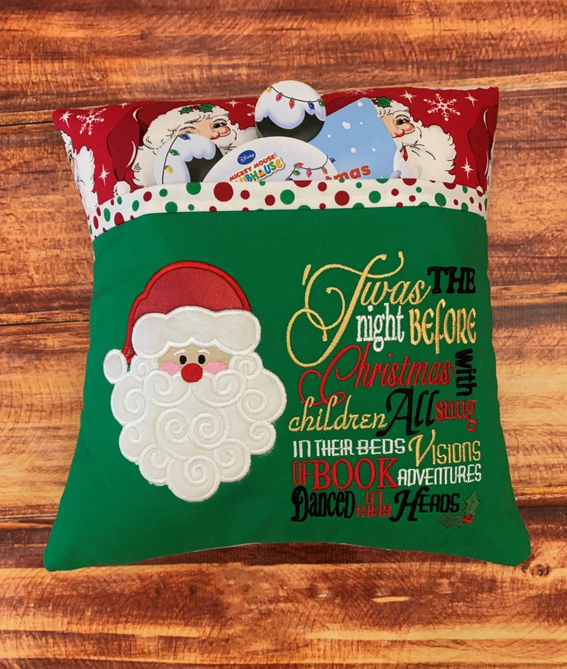 K&ouml;nnte beinhalten: Ein gr&uuml;nes Weihnachtskissen mit einem Santa Claus-Applikation und dem gestickten Text "Twas the night before Christmas". Das Kissen hat eine Tasche, die mit weihnachtlichen Gegenst&auml;nden gef&uuml;llt ist. Der obere Rand ist rot mit wei&szlig;en Punkten und Santa Claus-Illustrationen.