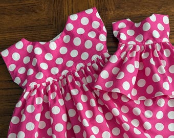 May include: Two pink dresses with white polka dots. The dresses have a sleeveless design with a gathered skirt. The fabric is a bright pink with large white polka dots. The dresses are displayed on a wooden surface.