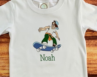 May include: White t-shirt with an embroidered image of a cartoon boy skateboarding. The boy is wearing a blue hat, green pants, and orange belt. The skateboard is blue and has black wheels. The name "Noah" is embroidered below the image.