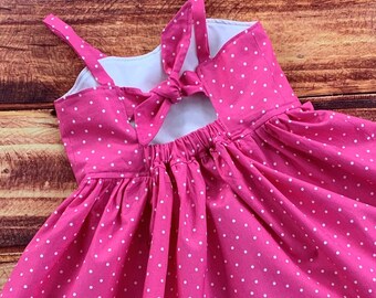 May include: A bright pink dress featuring a white polka dot pattern, thin shoulder straps, and a decorative bow detail at the center of the back. The garment includes a gathered skirt section and a white interior lining visible at the neckline.