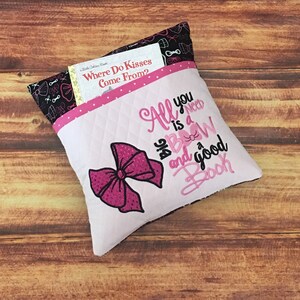 May include: A pink and white pillow with a large pink bow and the text "All you need is a bow and a good book". The pillow has a pocket on the back to hold a book.