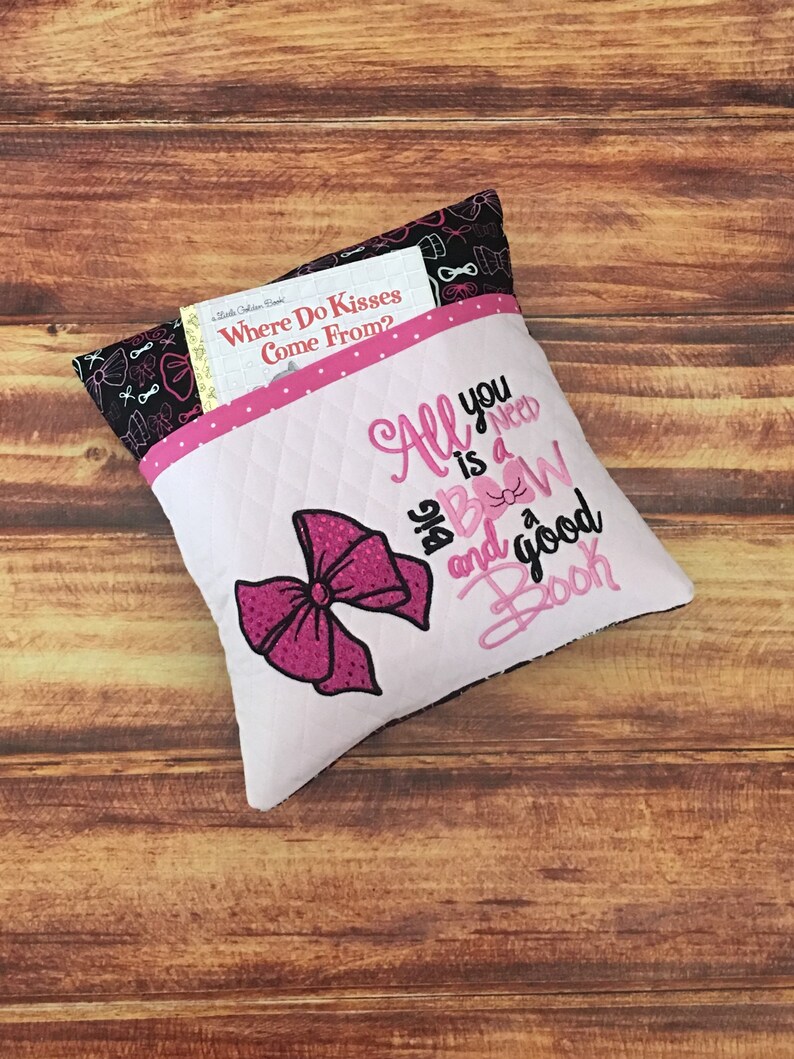 May include: A pink and white pillow with a large pink bow and the text "All you need is a bow and a good book". The pillow has a pocket on the back to hold a book.