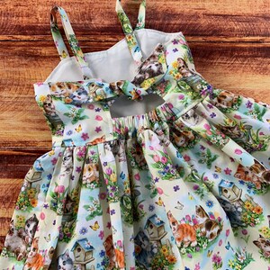 Pu&ograve; includere: Un abito senza maniche in tessuto chiaro con una stampa all-over di gattini, casette per uccelli, fiori colorati e farfalle. Il capo presenta spalline sottili, un dettaglio da annodare sul retro e una gonna arricciata e pieghettata.