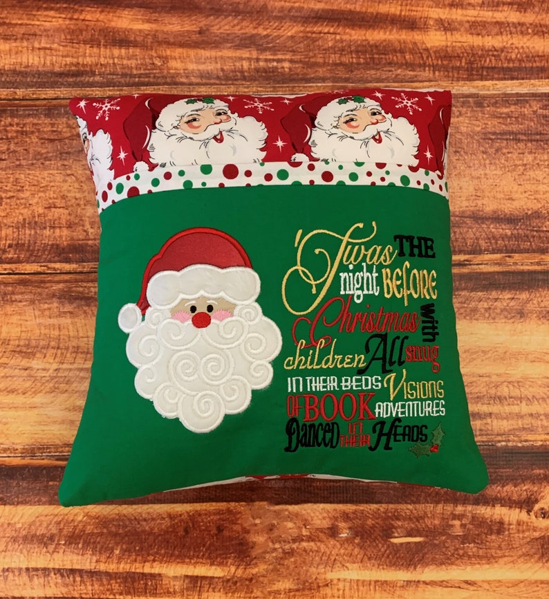 K&ouml;nnte beinhalten: Ein quadratisches Weihnachtskissen mit einer gr&uuml;nen Vorderseite, auf der ein gestickter Weihnachtsmann und der Text "Twas the night before Christmas" zu sehen sind. Der obere Rand ist rot mit Weihnachtsmann-Gesichtern und Schneeflocken.