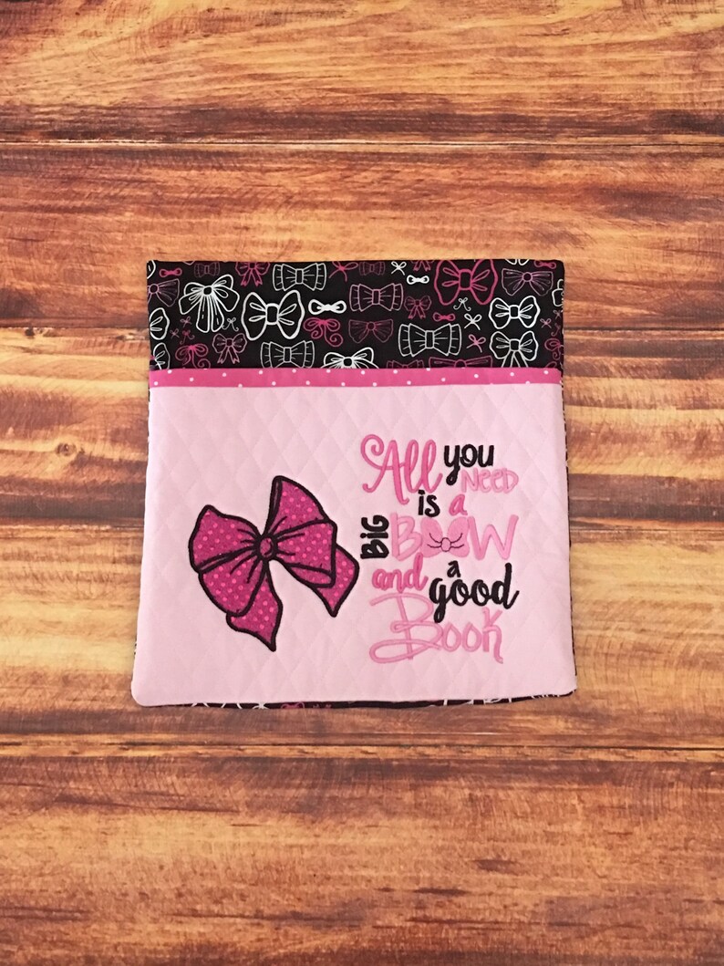 May include: Pink fabric pouch with a black and white bow pattern on the back. The front of the pouch is pink with a large pink bow and the text "All you need is a big bow and a good book".