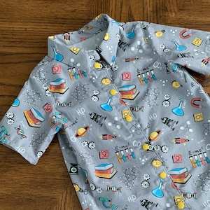 May include: A gray short-sleeved button-down shirt with a science theme. The shirt has a variety of science-related images, including test tubes, molecules, and a rocket ship. The text "E=mc²" and "H₂O" are printed on the shirt.